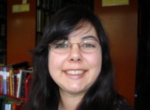 Image of Kiyomi Deards head with bookcases behind her. Kiyomi is a Japanese American female with long dark hair, bangs, and glasses. She is smiling and wearing mauve lipstick.