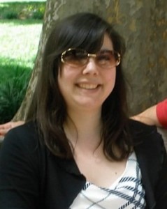 Image of Kiyomi Deards a Japanese American Woman with long dark brown hair, bangs, and gold rimmed brown tinted sunglasses in from of a tree with a black sweater and white dress with blue lines.