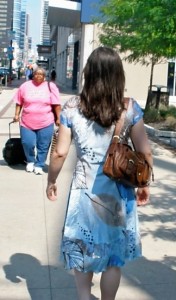 Image of a woman walking away down the street with long dark hair, wearing a blue midi dress and a brown shoulderbag.