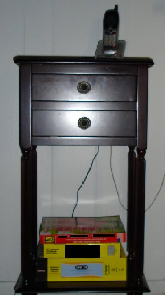 Photo of a black wireless phone handset on top of a dark cherry wood phone stand against a white wall.