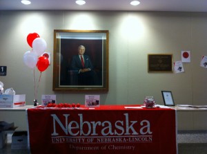 Image of Hamilton Hall featuring a portrait of Dr. Hamilton in the background with a table in the foreground containing red and white ballons and a red chemistry department table cloth for UNL Chemistry Department on Chemistry Day at UNL.