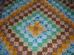 Image of a Quilt made by Jodye Selco showing a progressing diamond pattern of block starting with yellows and browns and progressing to blues and blue green, in a repeating pattern.