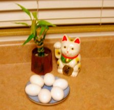 Photo of a small blue plate with six white eggs in front of a small bamboo plant and white Japanese lucky cat on a light brown counter.