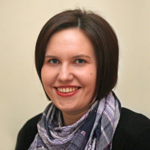 Picture of Jo Alcock. Jo has light skin with short brown hair and is wearing a blue and pink plaid scarf, black top, and a medium dark pink lipstick.
